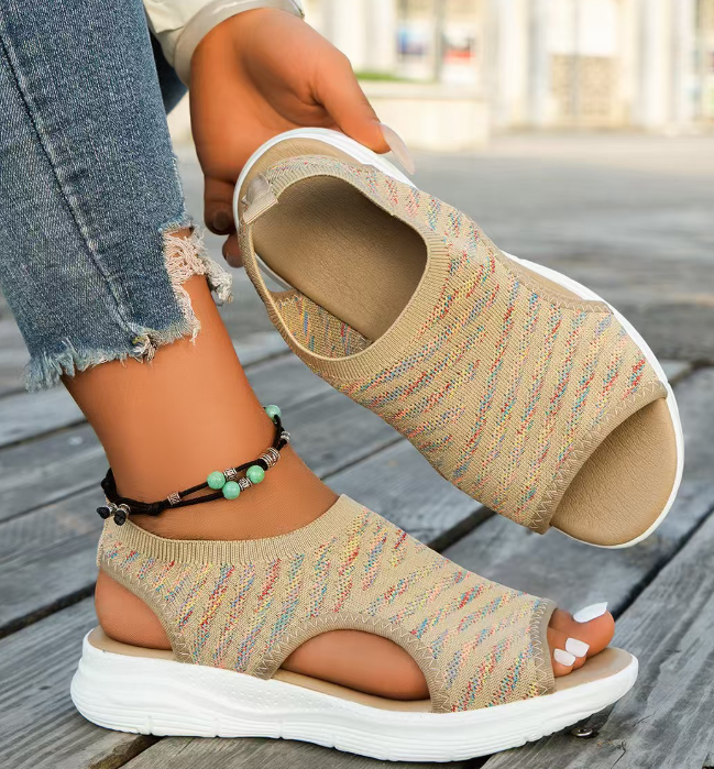 Beige open-toe sandals with white soles worn by a person on a wooden floor.